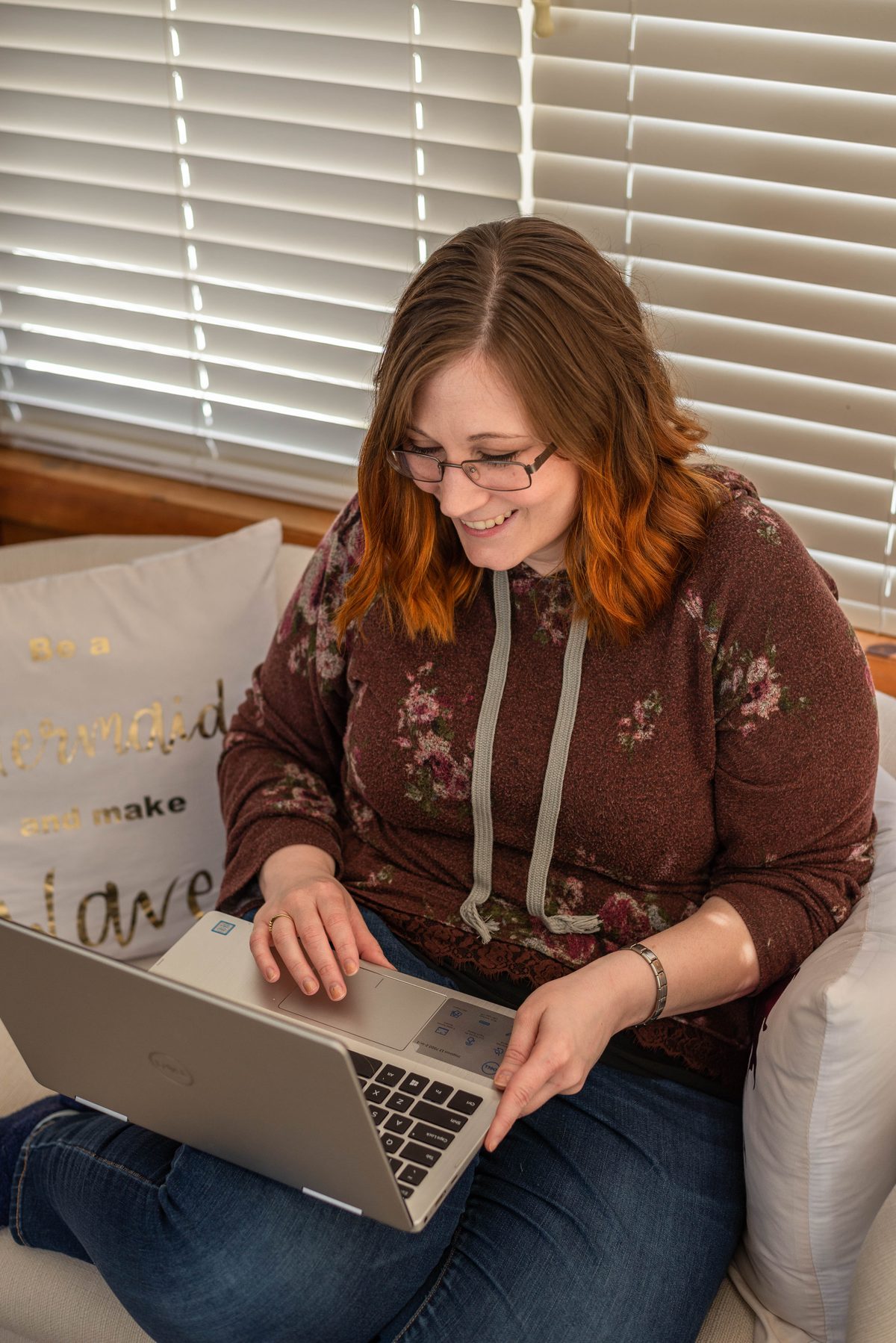 Ariel smiling down at a laptop balanced on her knees as she works from a couch by a sunlit window.