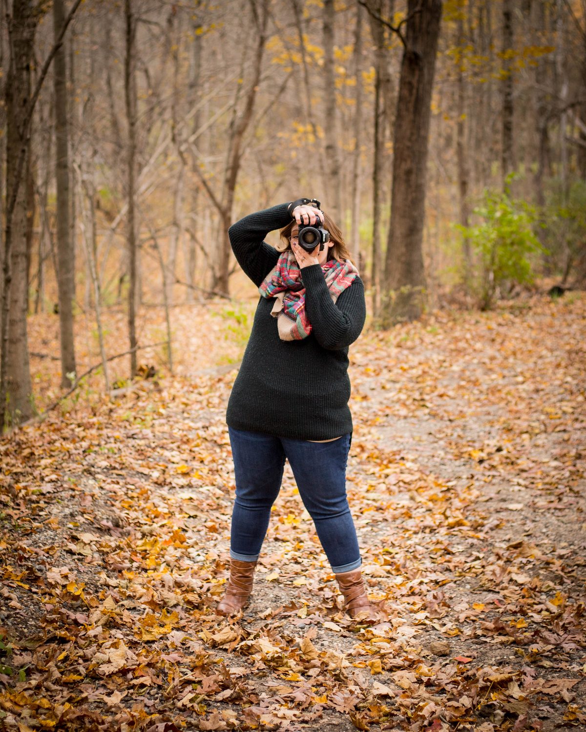 Ariel raising a camera to her eye on a leaf-covered path in an autumn woods, in a dark sweater and plaid scarf.