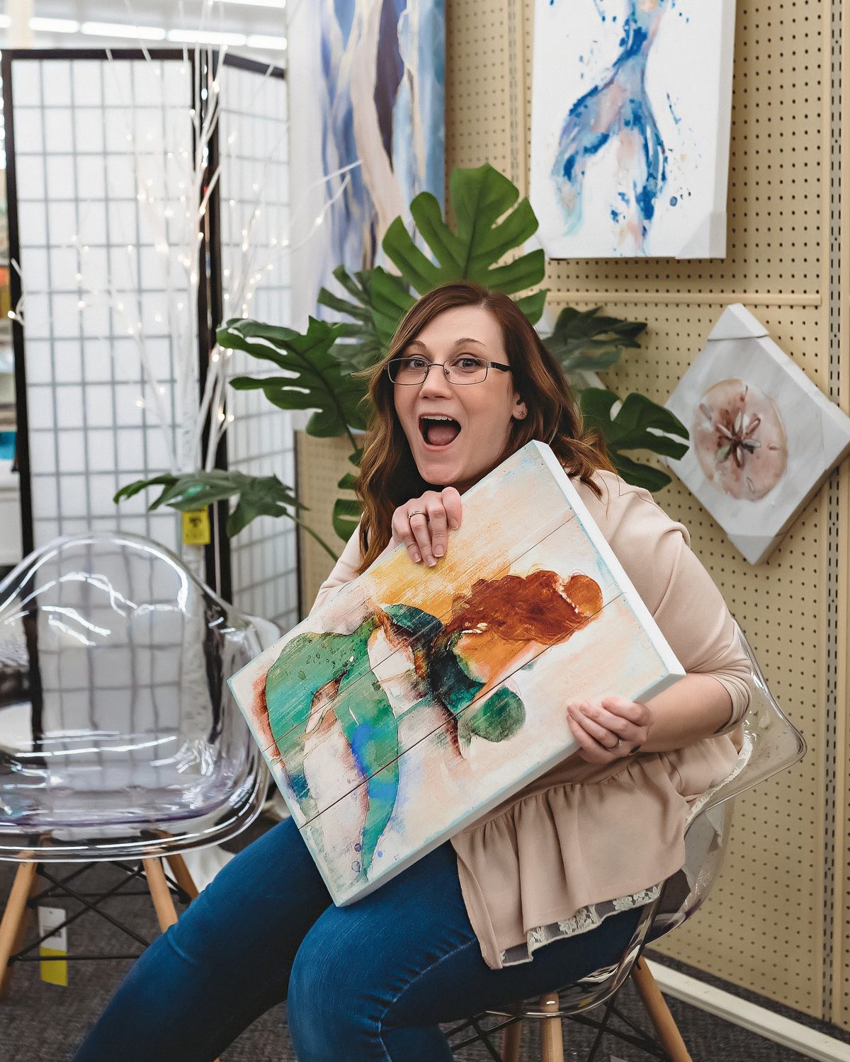 Ariel with a wide, delighted expression, holding a colorful mermaid painting on a wood panel in a shop full of artwork.