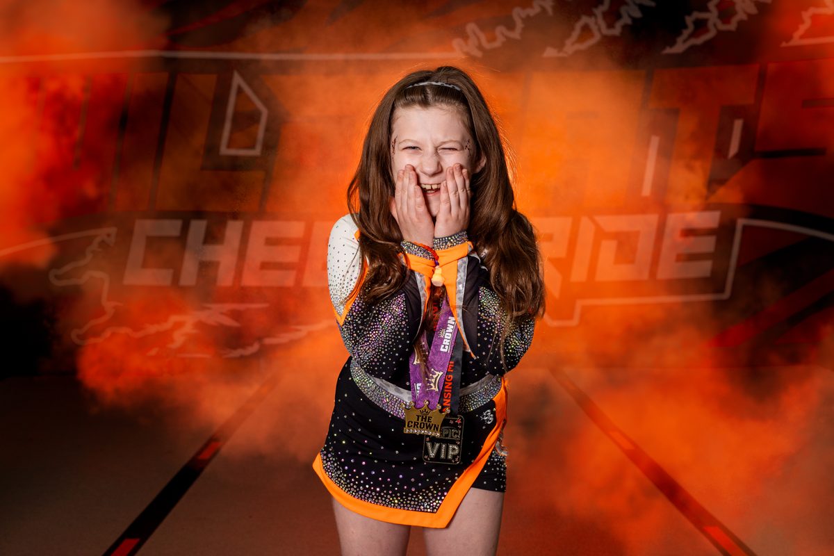 A young competitive cheerleader laughing with her hands to her cheeks, in a rhinestone uniform and competition medals, surrounded by orange smoke.