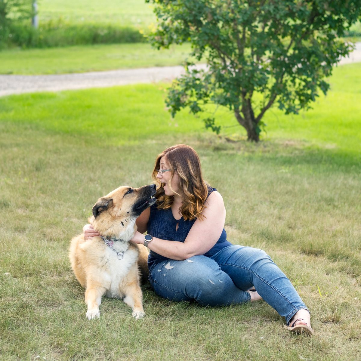 Ariel sitting on a lawn and laughing as her dog leans in to lick her face.
