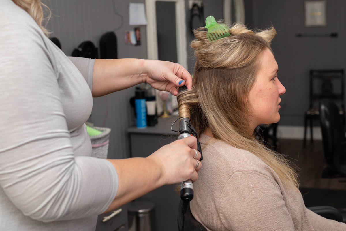 A hairstylist wraps a section of a client's hair around a curling iron in a salon.