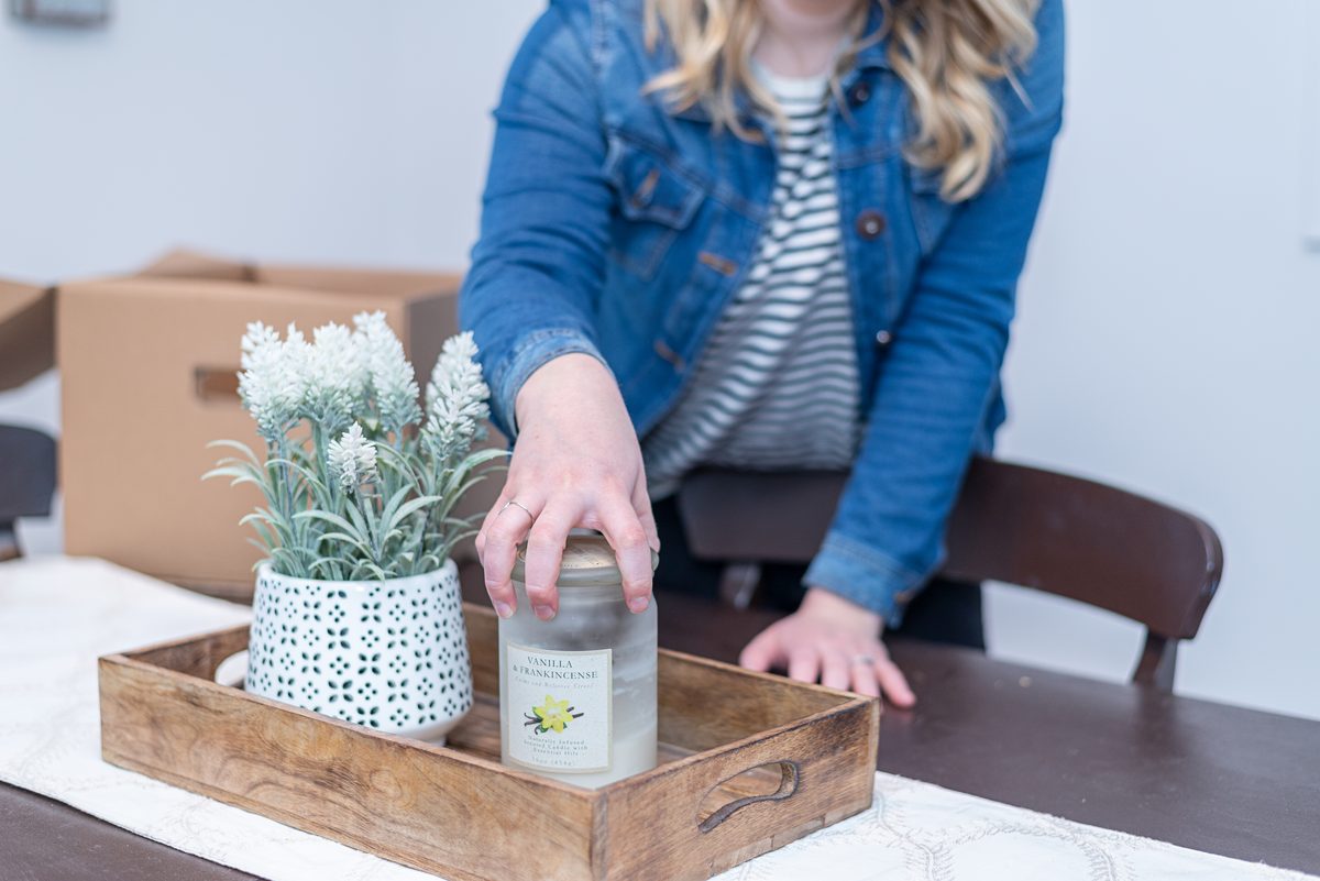 A woman in a denim jacket sets a glass jar candle with a lemon label on a wooden tray beside a potted white flower.