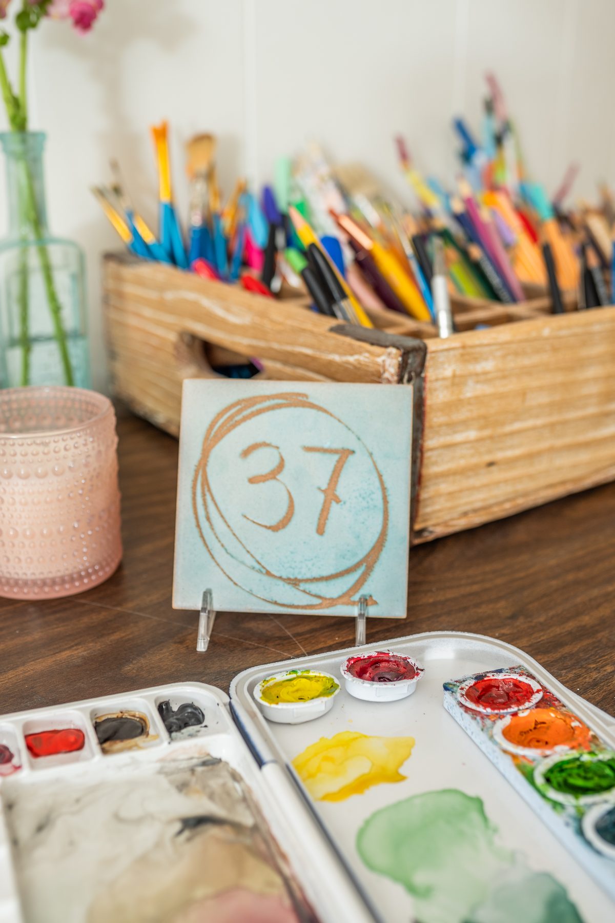 A wooden crate of paintbrushes and pencils, a hand-painted number 37 tile and a watercolor palette on a wooden table.