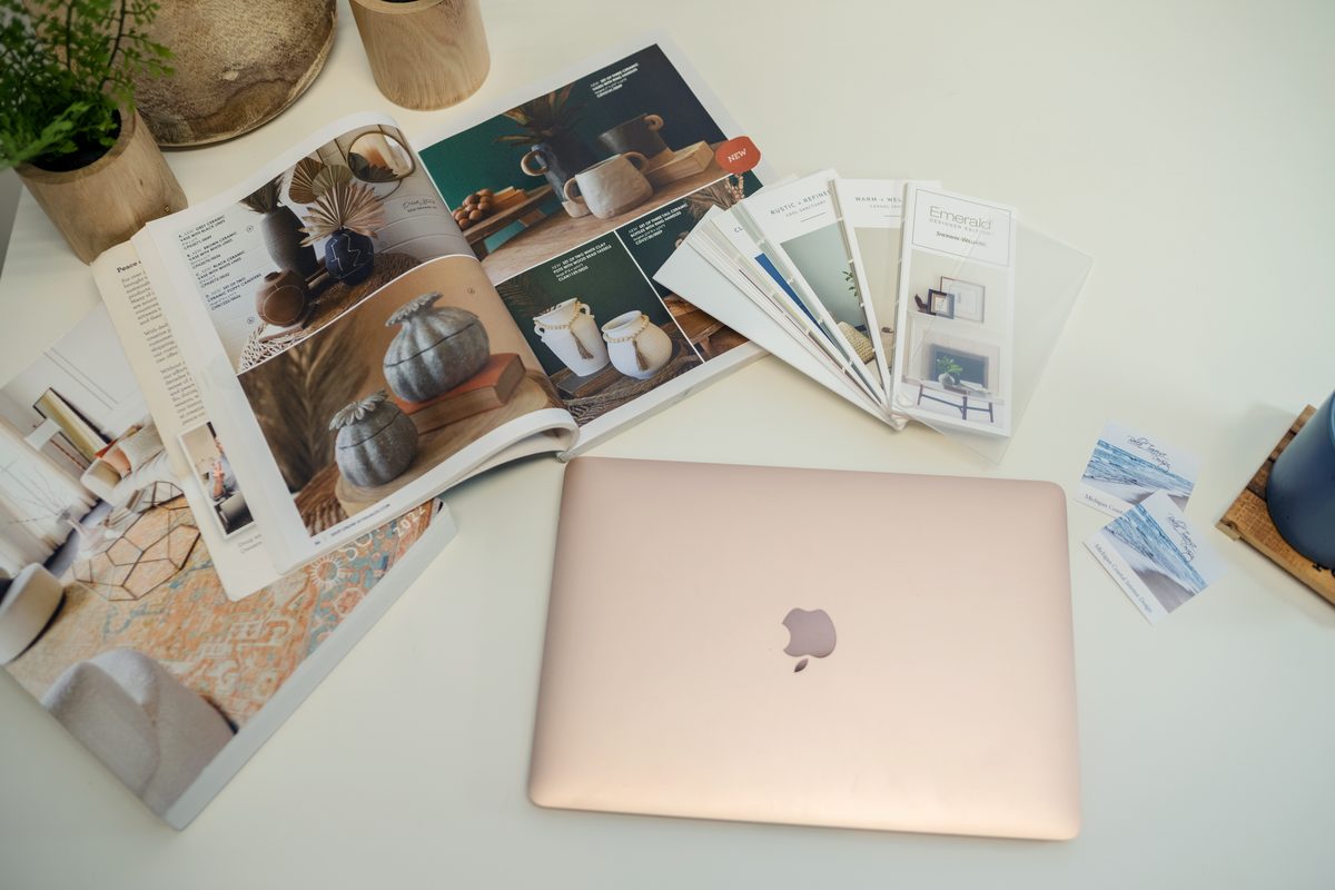 An overhead view of a white table with an open home-decor catalog, paint and color brochures, swatches and a rose-gold laptop, with a potted plant at the edge.