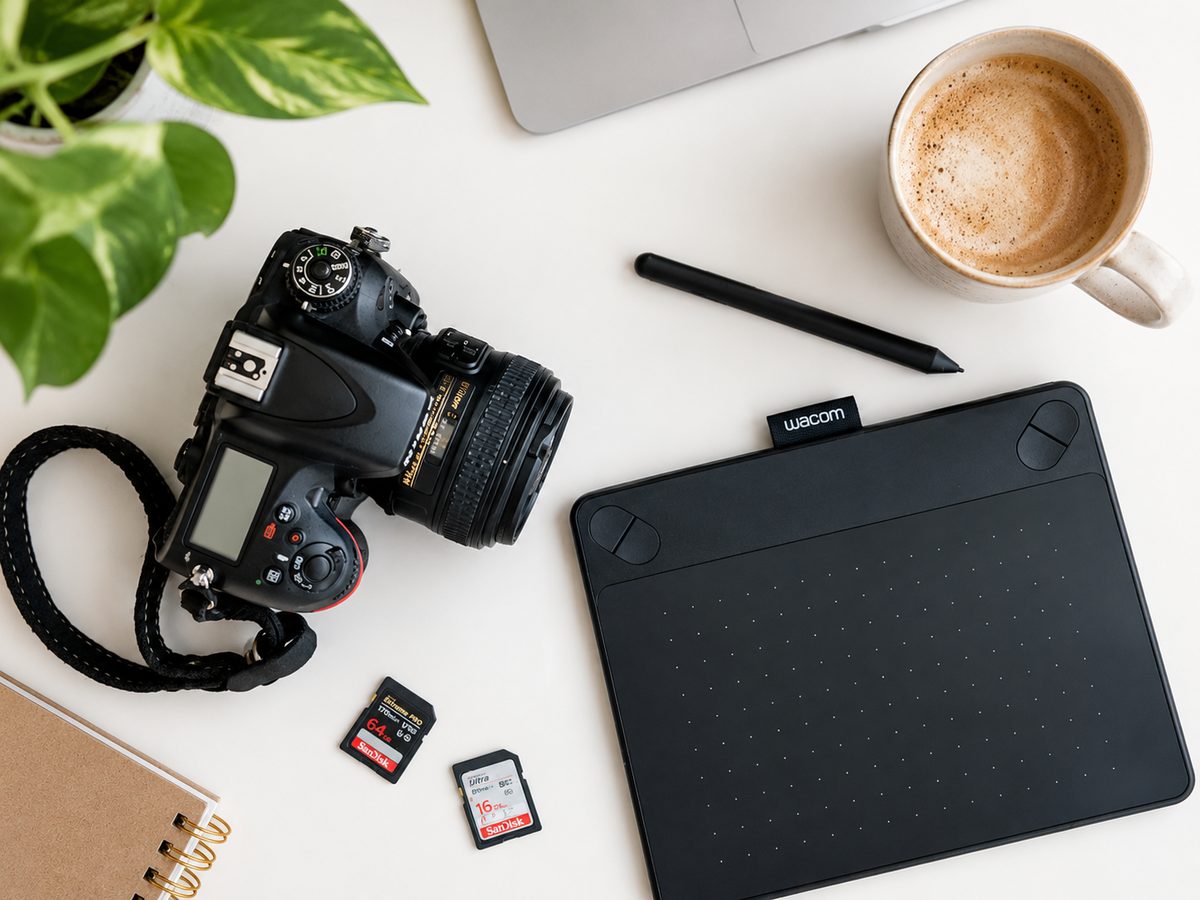 An overhead desk with a camera, a black drawing tablet and pen, two memory cards, a notebook and a cup of coffee beside a trailing plant.