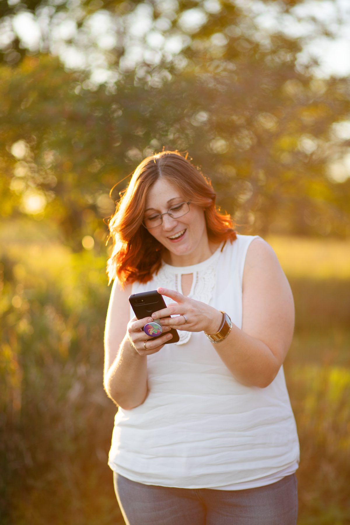 A smiling woman with red hair and glasses in a white top looks down at her phone in a sunlit field at golden hour.