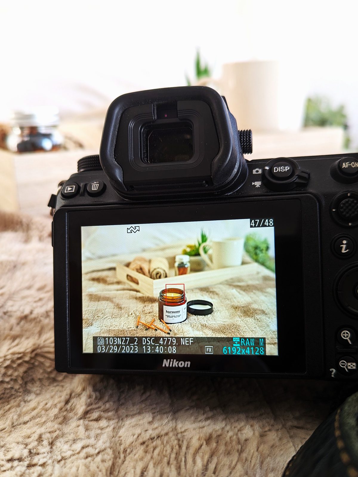 The back of a camera showing a photo of a jar candle on a tray in its screen, resting on a soft fur blanket.