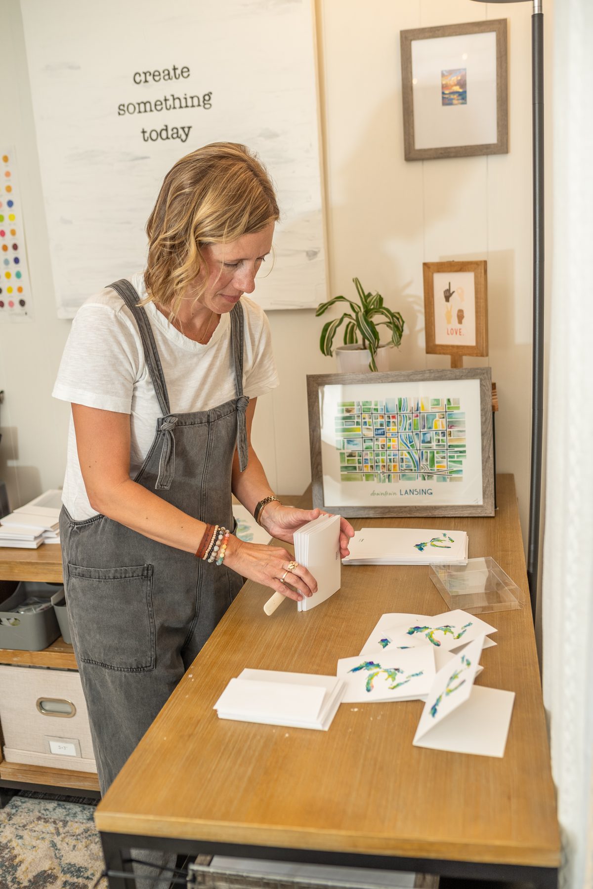 A woman in a white tee and gray overalls arranges hand-painted watercolor cards on a wooden table in a bright studio with framed prints and a plant behind her.