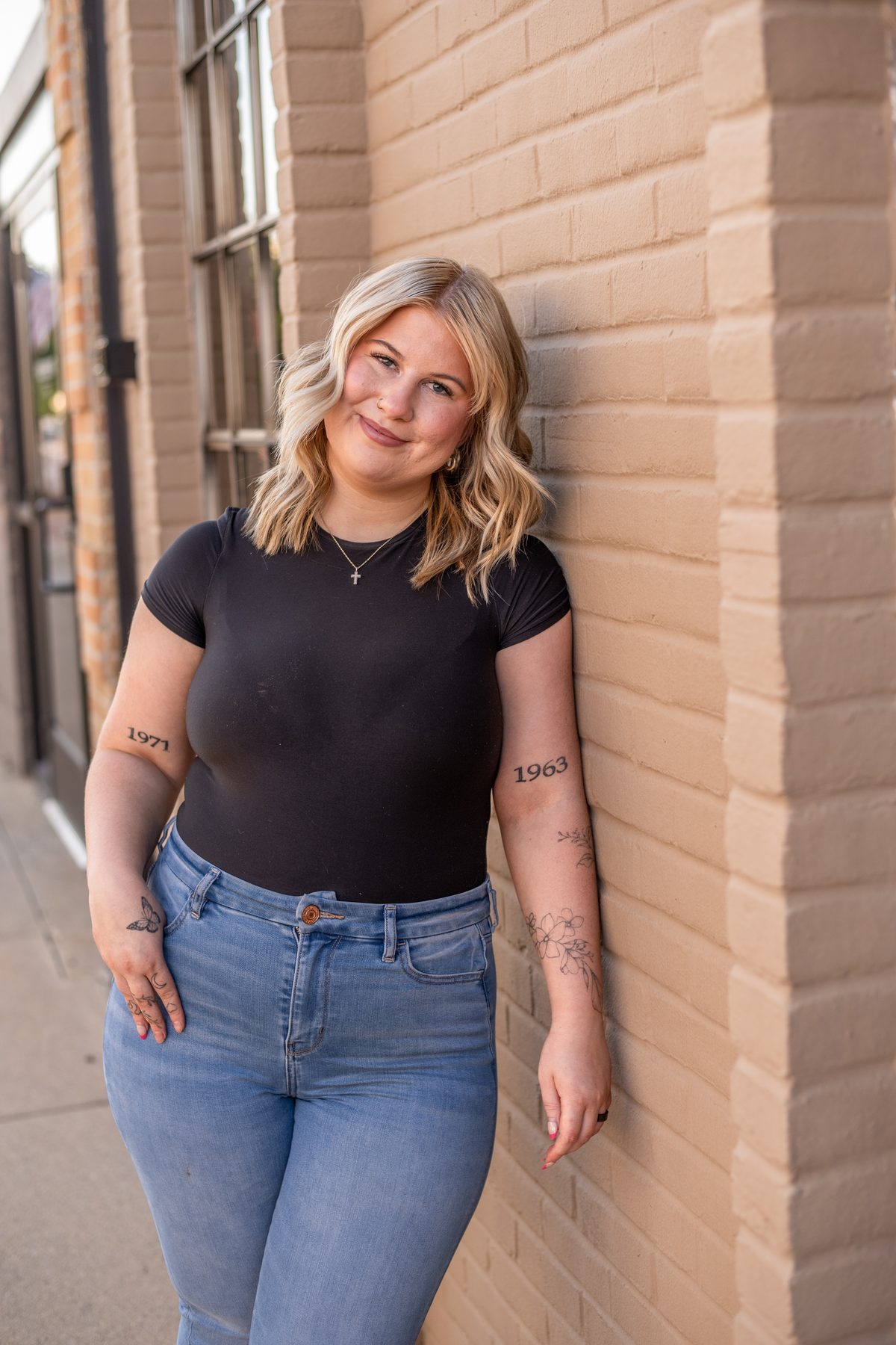 A smiling woman with wavy blonde hair in a black tee and jeans leans against a peach-colored brick wall, with tattoos on her arms.