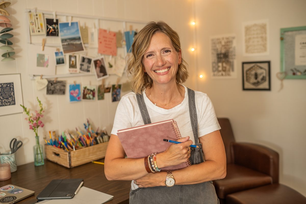A smiling woman in a white tee and gray overalls holds a pink notebook and pen in front of a studio wall of pinned photos and string lights.