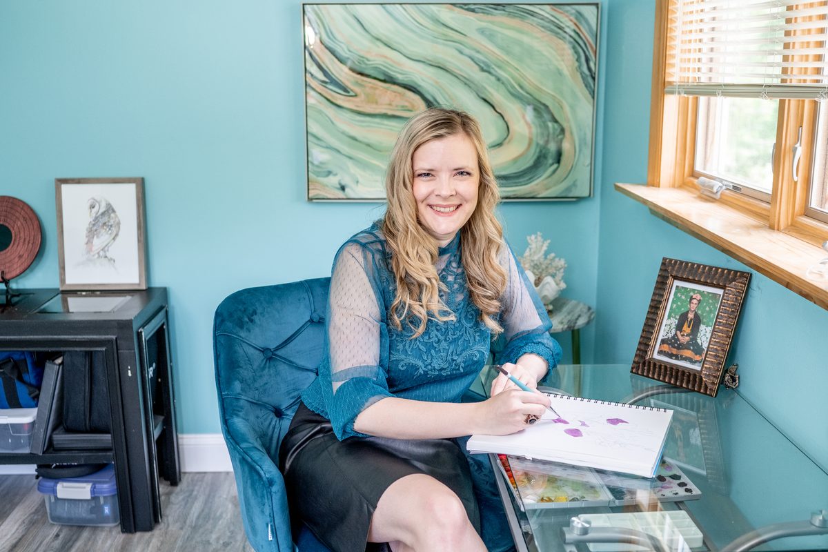 A woman in a teal lace blouse sits at a glass desk in a teal-walled studio, painting in a sketchbook.