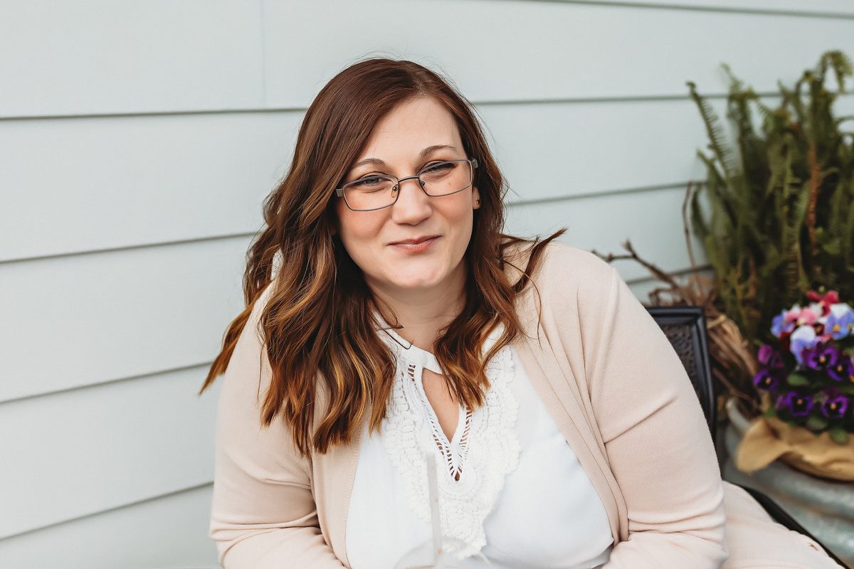 A smiling woman with glasses and long auburn hair, in a cream cardigan, seated outdoors in front of pale siding and a planter of pansies.