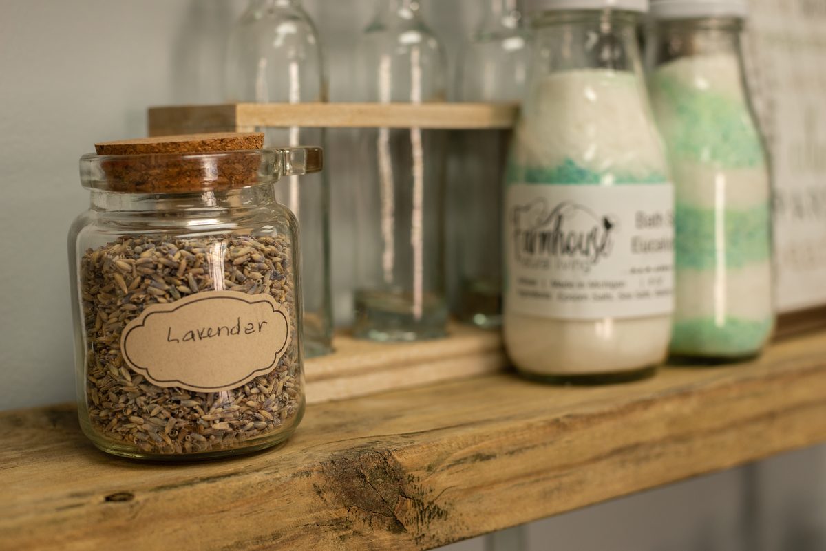 A glass jar of dried lavender with a handwritten label on a rustic wooden shelf beside bottles of layered bath salts.