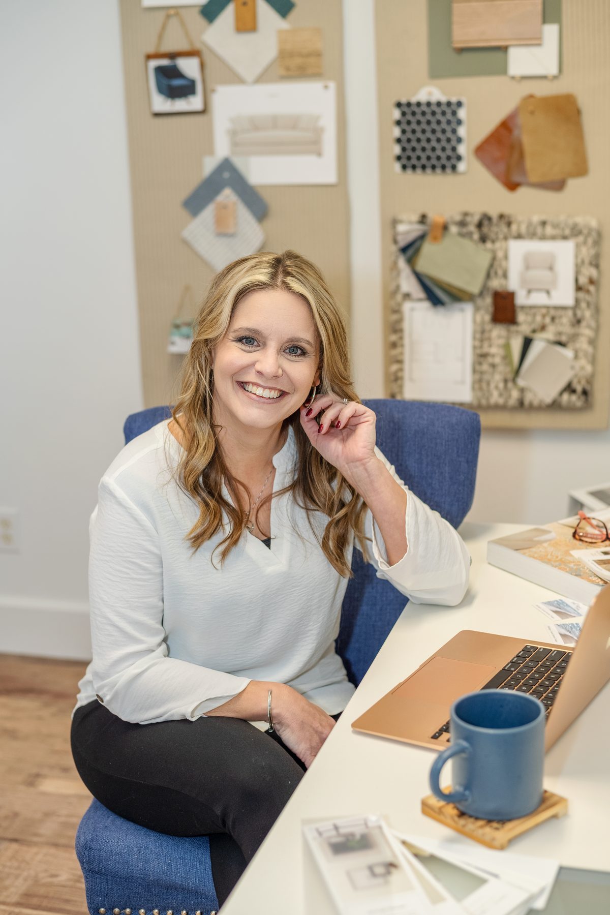 A smiling interior designer seated at her desk beside a laptop and a blue mug, with fabric and material samples pinned to the boards behind her.