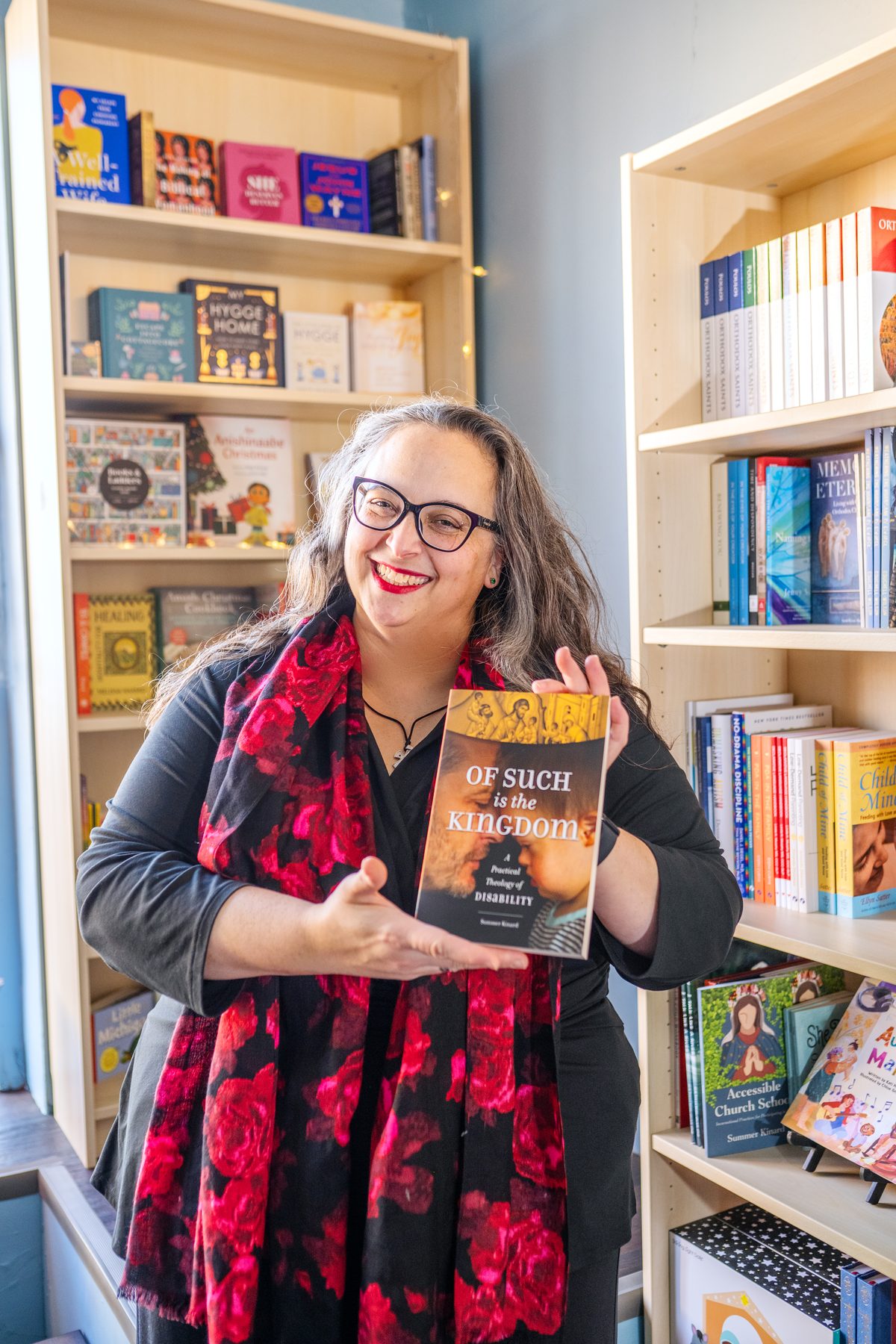 A smiling woman in a red floral scarf holding up a book in front of crowded bookshelves.