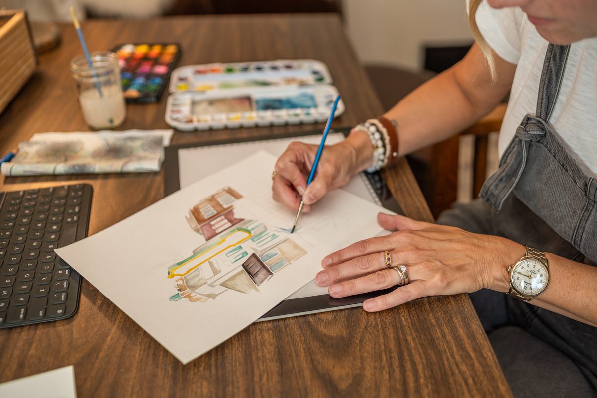 Hands painting a watercolor of a building at a wooden desk, with a paint palette and brushes close by.