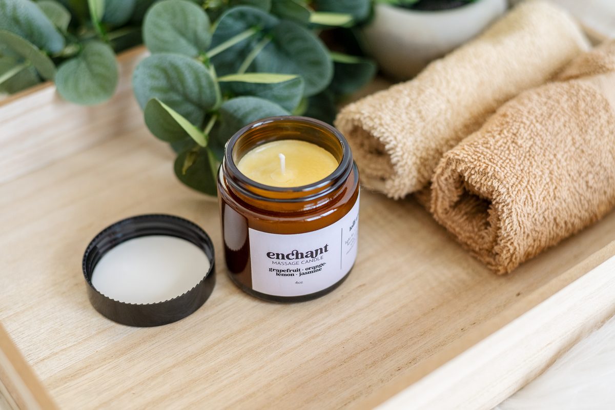 An amber jar candle with its lid set beside it on a wooden tray, with eucalyptus leaves and rolled towels.