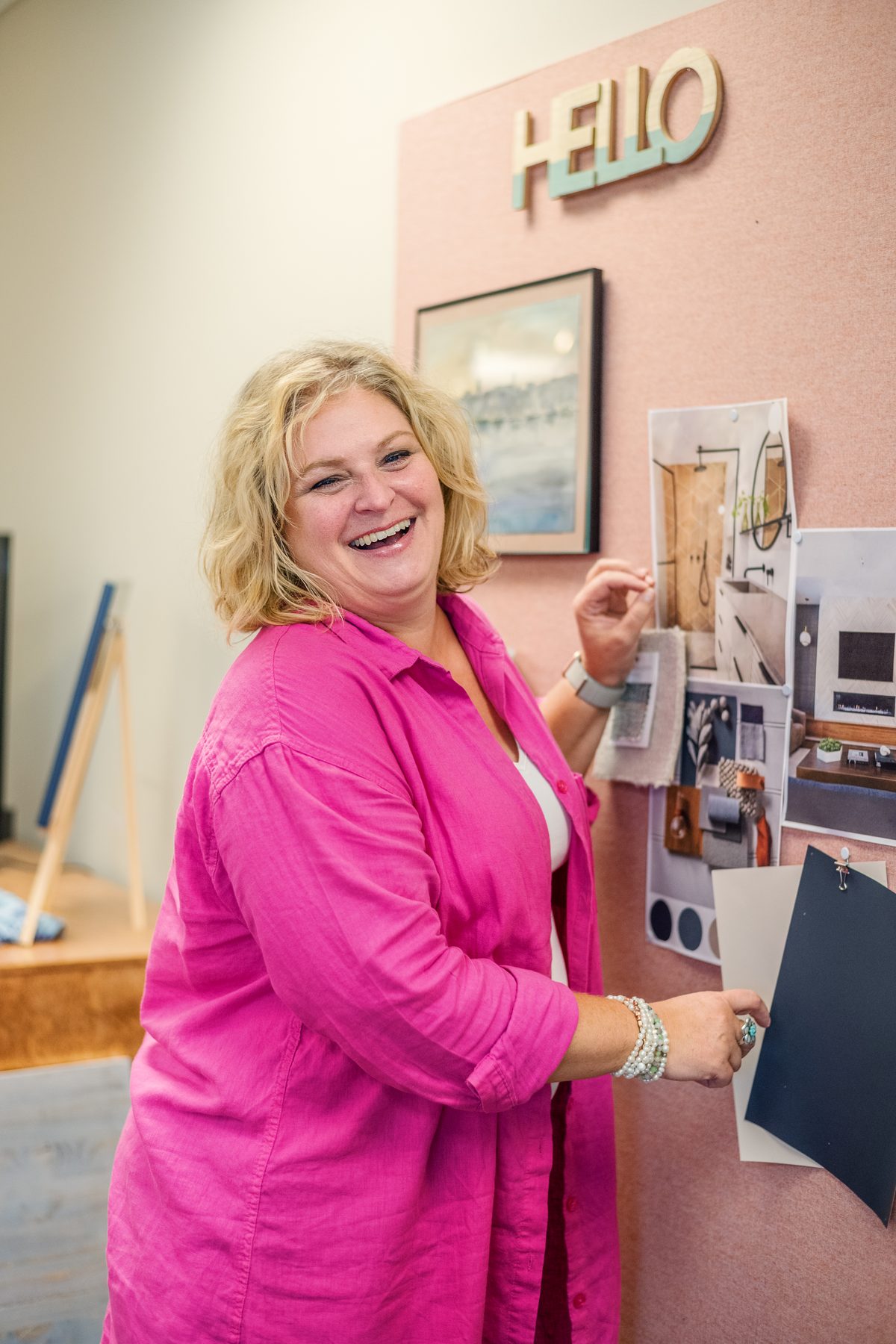 A laughing woman in a bright pink shirt placing a fabric sample on a pink mood board beneath a HELLO sign.