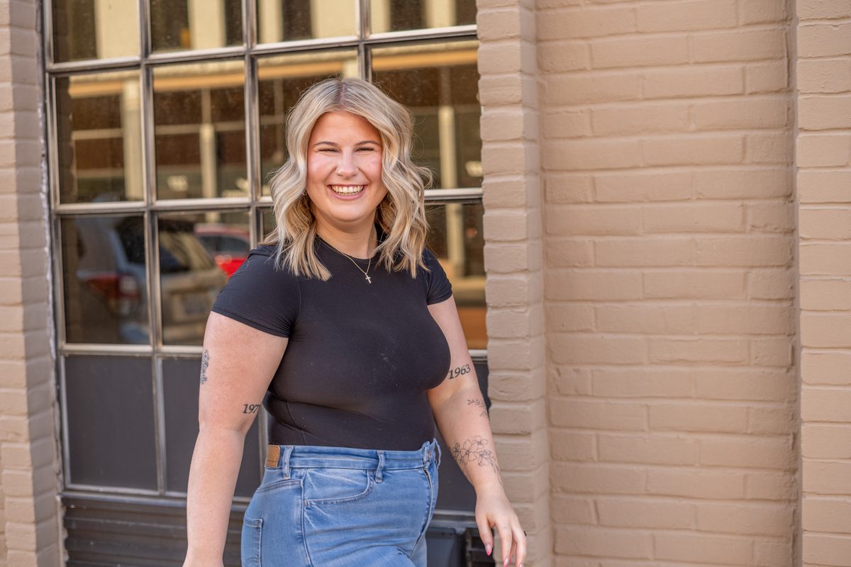 A woman with blonde wavy hair laughing outdoors in a black top and jeans, in front of a brick storefront window.