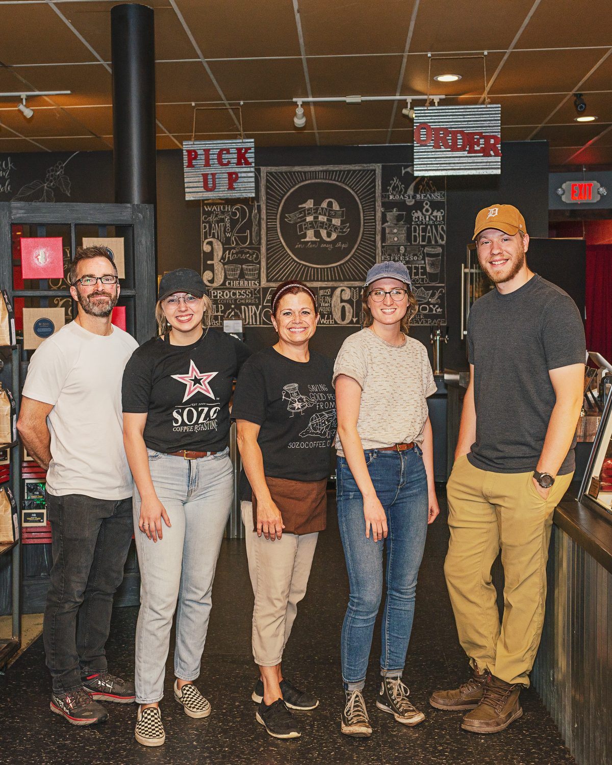 Five coffee roastery team members smiling together in front of a chalkboard wall and Pick Up and Order signs.