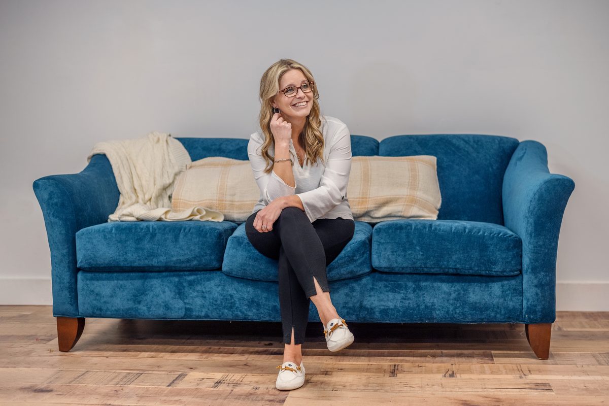 A smiling woman with glasses seated on a teal velvet sofa in a bright studio, one hand raised to her ear and her legs crossed.