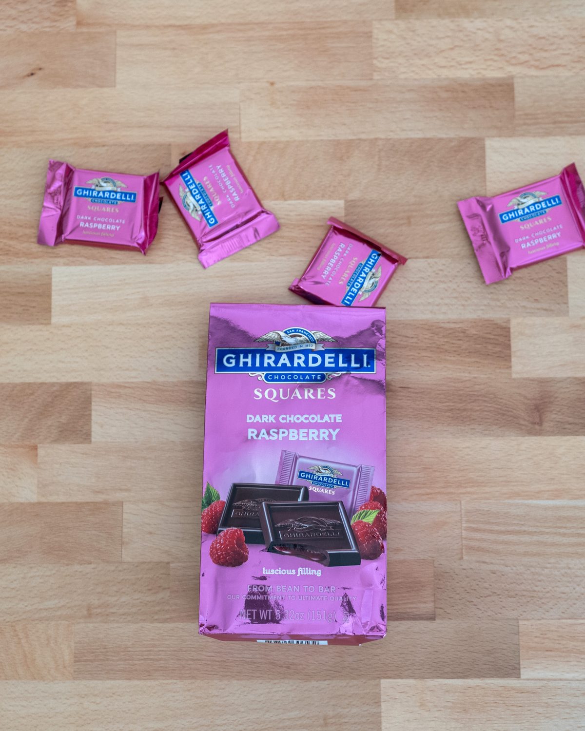 A pink Ghirardelli dark chocolate raspberry bag stands on a light wood table with four wrapped squares scattered beside it, one still sliding out of the bag.