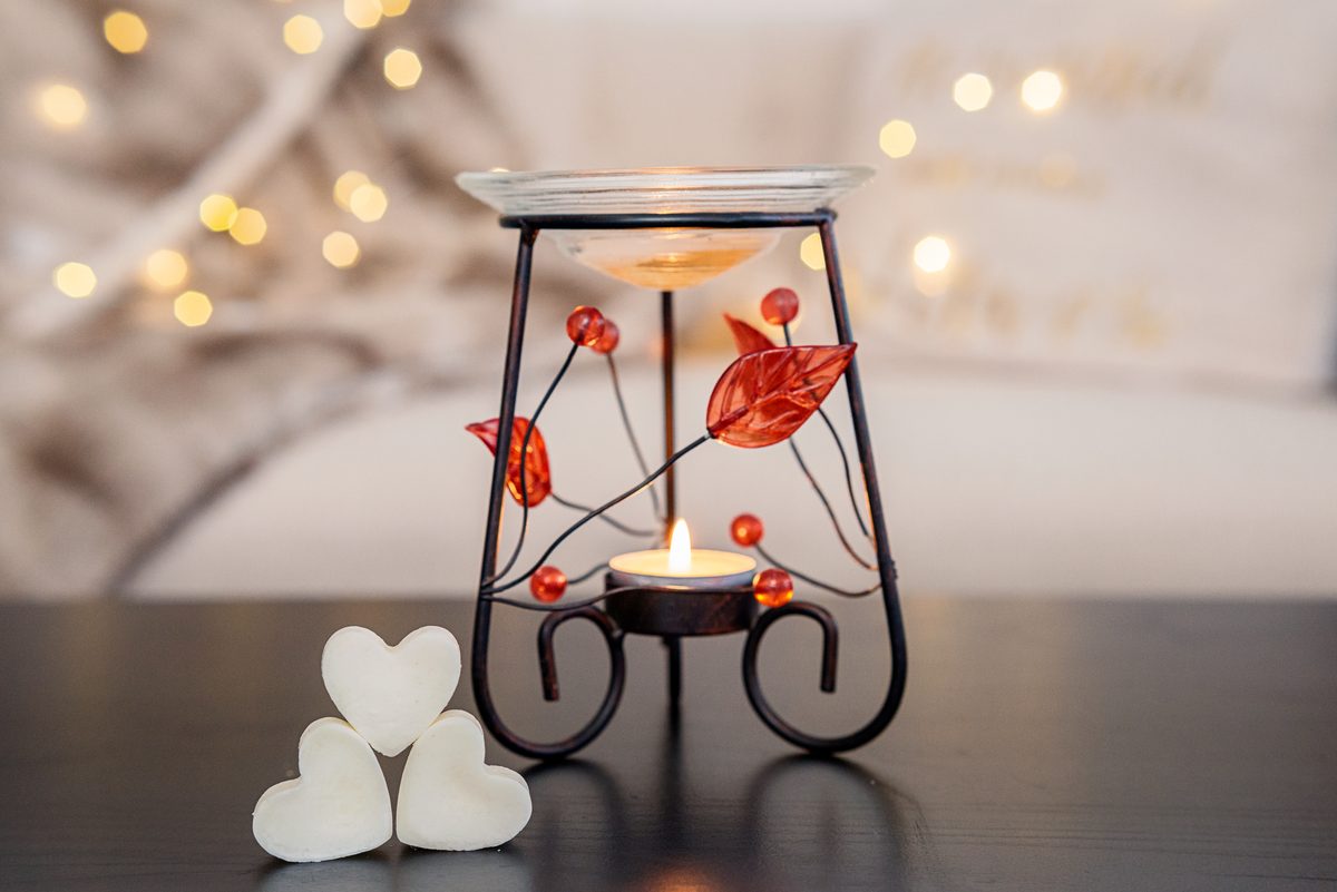 A black wire oil warmer with red glass leaves and beads holds a lit tea light under a glass dish, with three white heart-shaped wax melts beside it on a dark table and warm lights glowing behind.