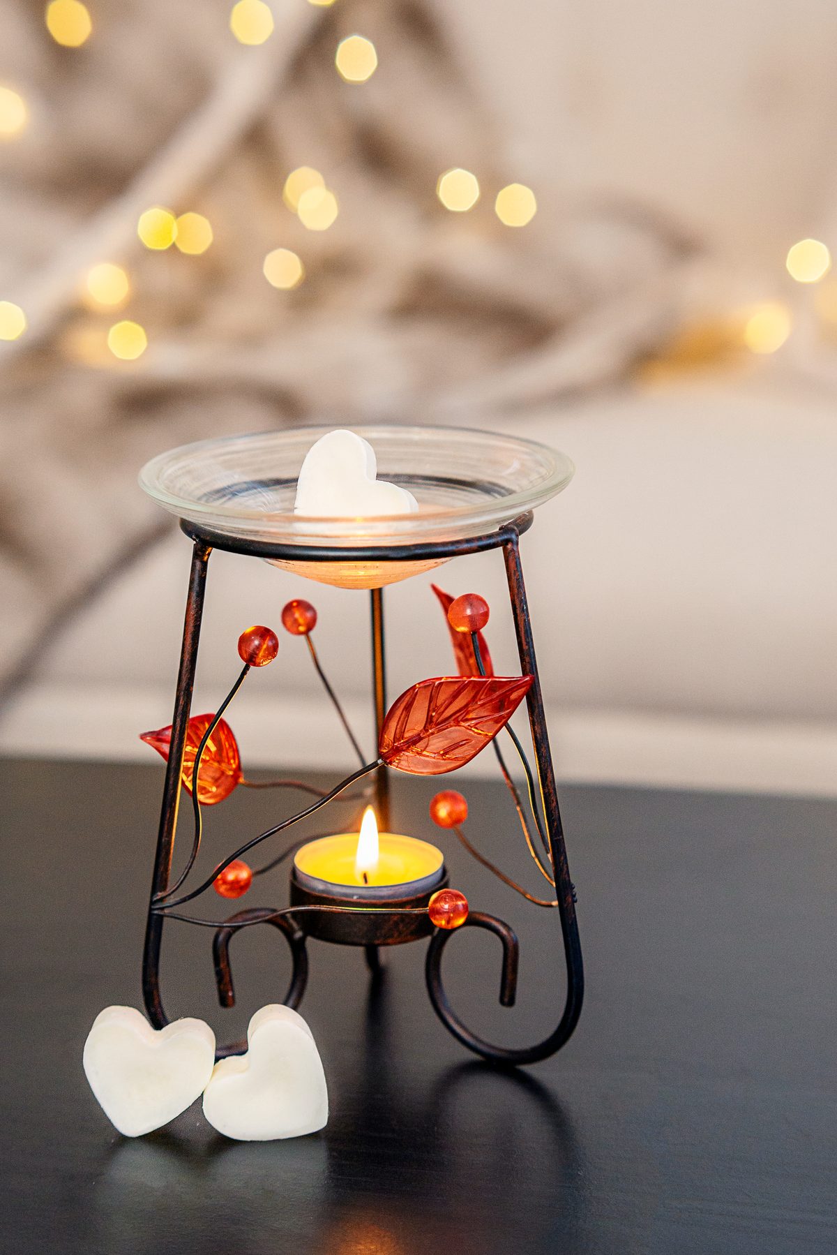 The same oil warmer with a white heart-shaped wax melt resting in its glass dish above a lit tea light, with two more hearts at its base and soft string lights behind.