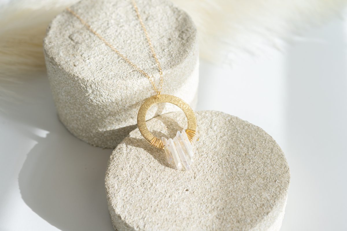 A gold circle necklace with clear quartz crystals resting on rough, sand-colored stone in soft window light.