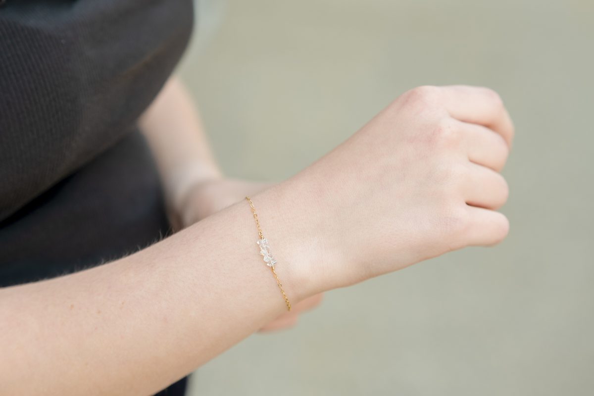 A closed hand and wrist wearing a thin gold bracelet with small clear crystals, against a soft grey-green background.