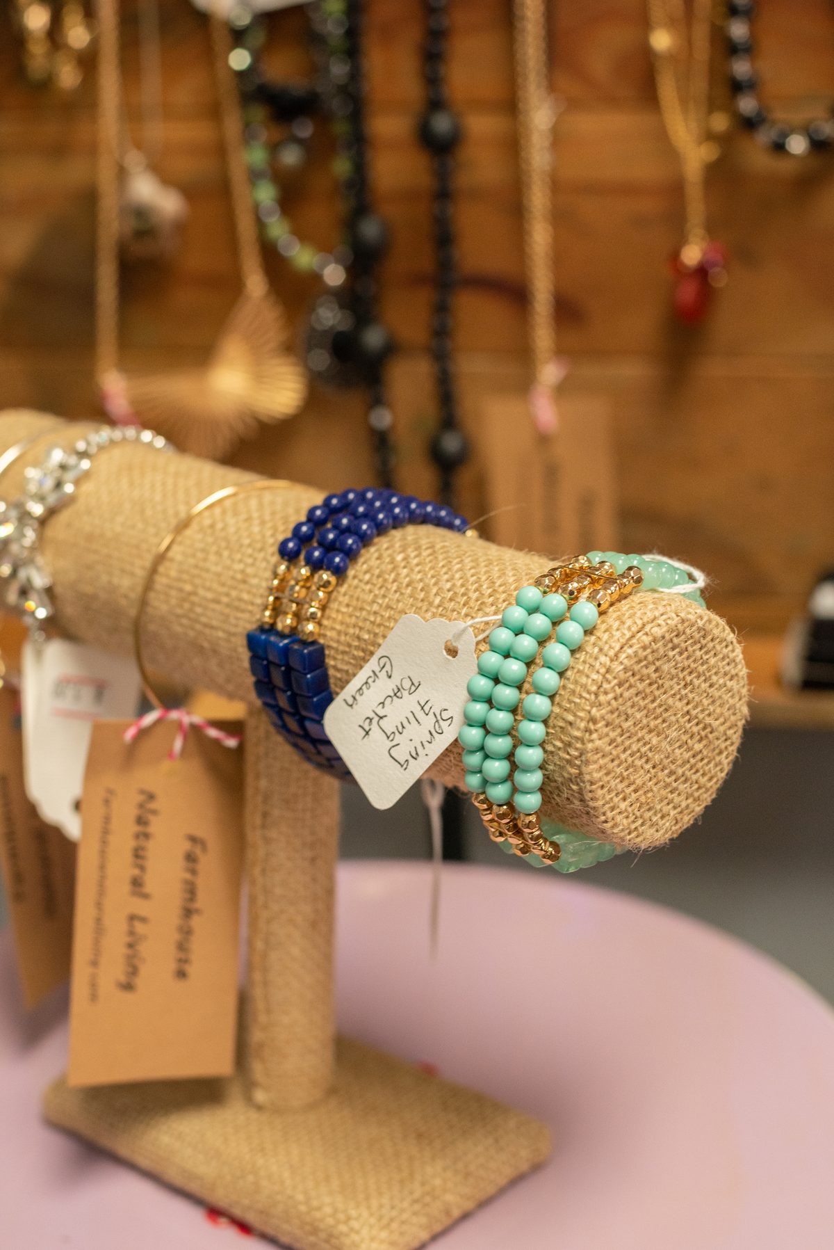 A burlap jewelry stand holding blue and mint-green beaded bracelets with handwritten tags, with necklaces hanging on the wall behind.