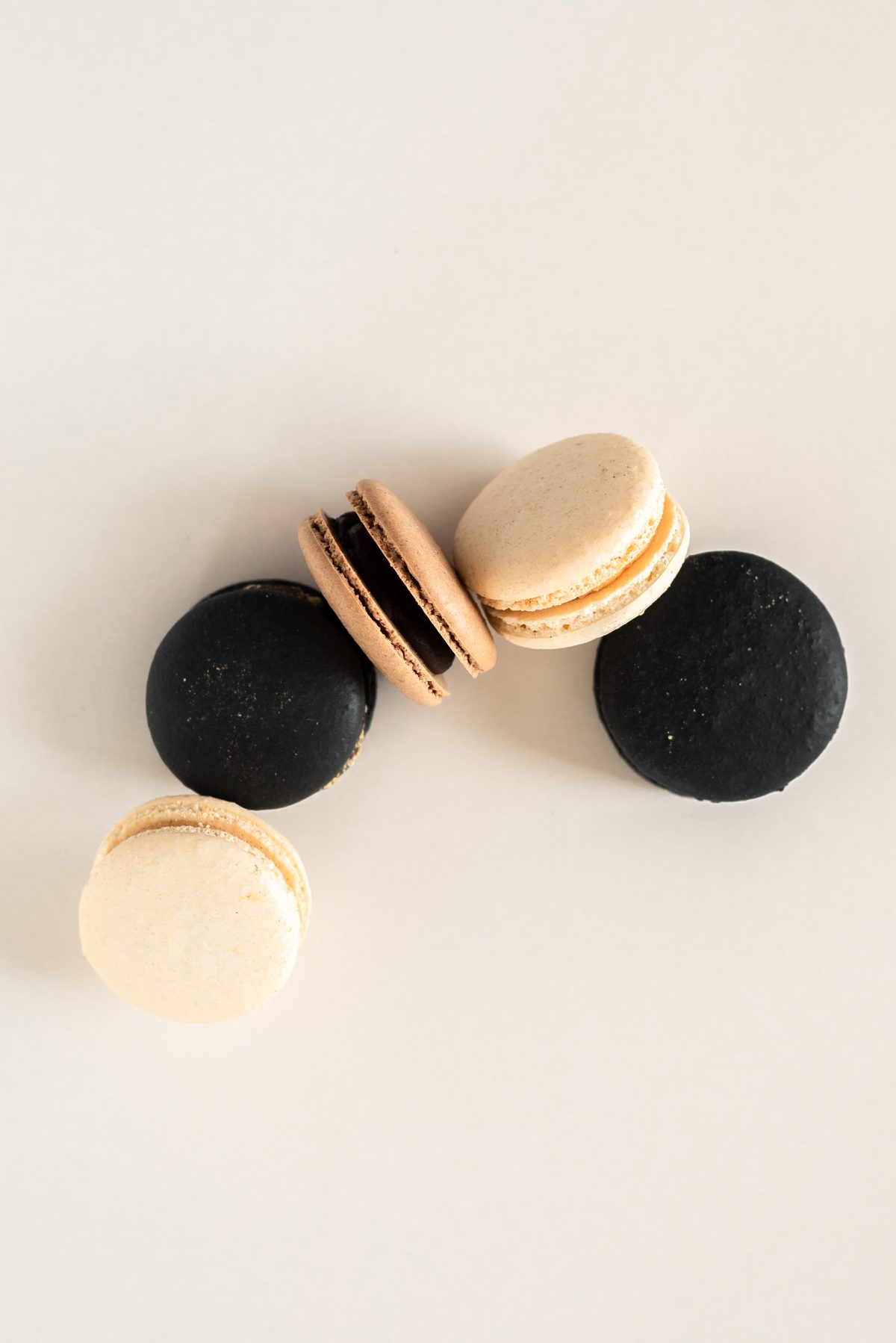 Five macarons in black, tan and cream scattered across a plain cream surface.