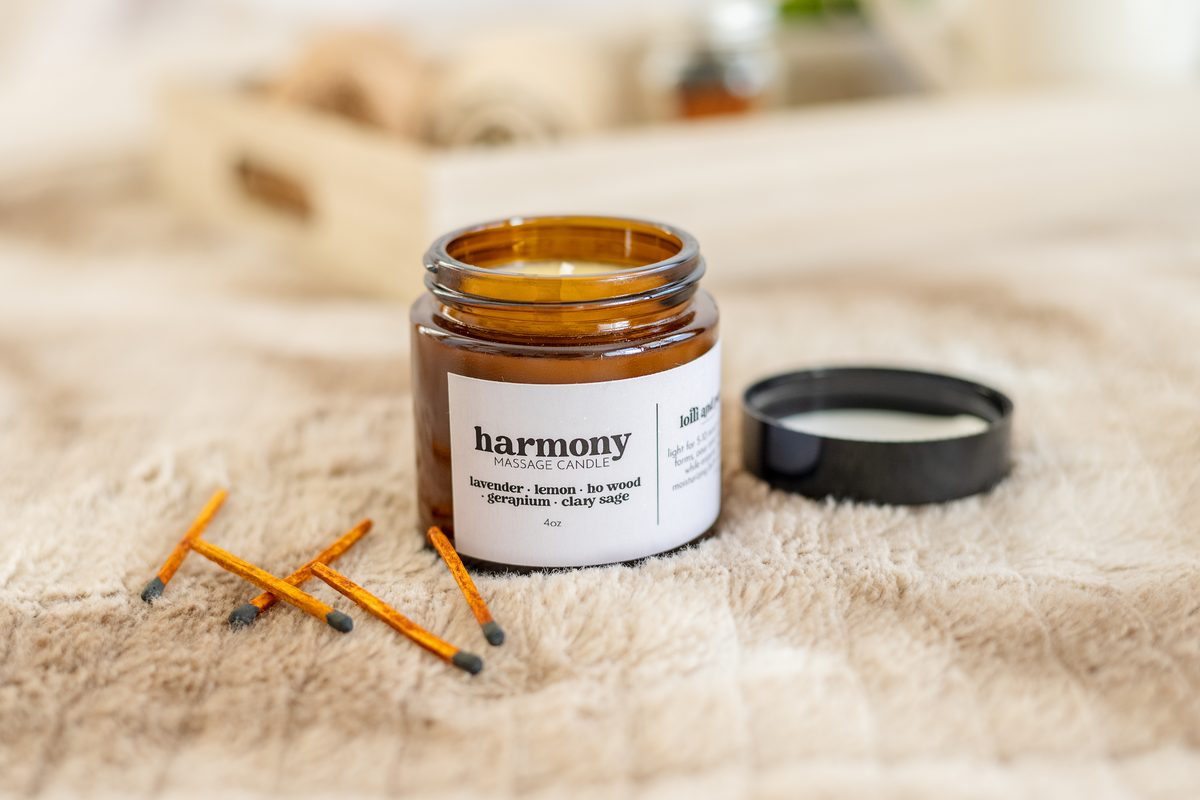 An amber jar candle with its lid set beside it and orange-tipped matches scattered across a soft cream blanket, with a wooden tray behind.