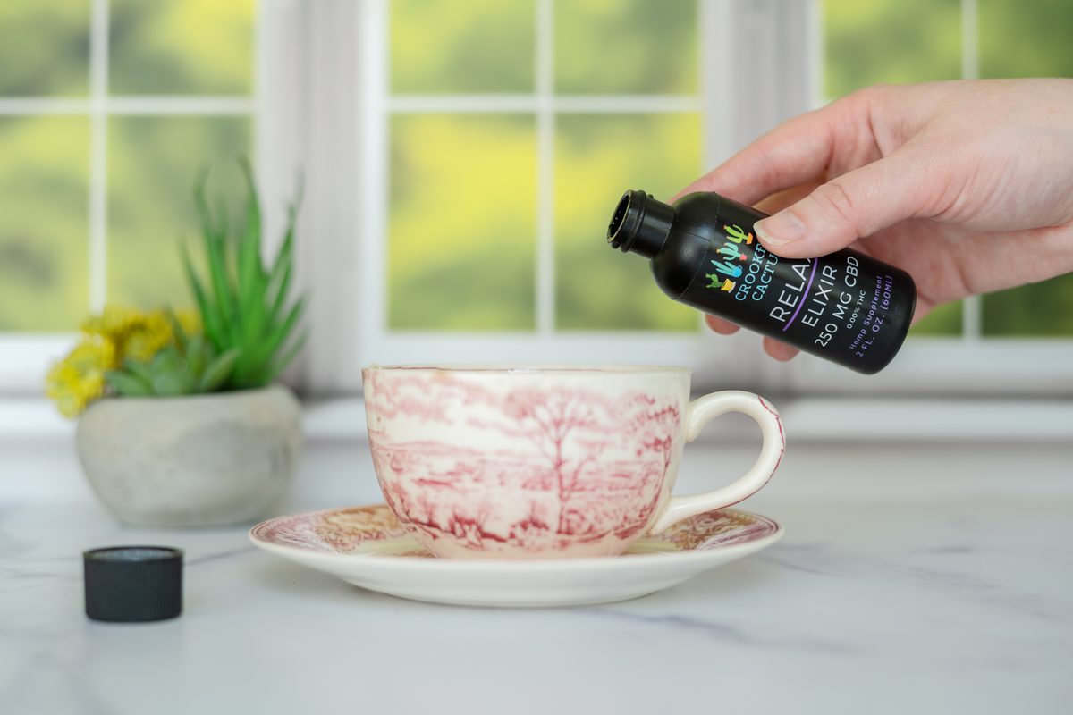 A hand tipping a black dropper bottle of elixir toward a pink-and-white teacup on a marble counter in front of a bright window.