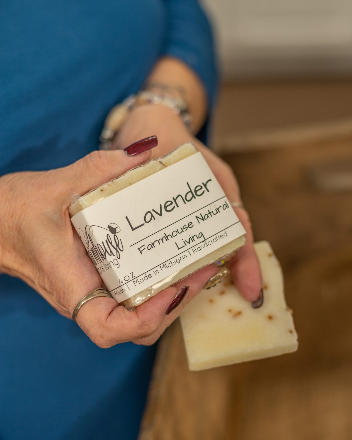 A woman in a teal top holding a bar of lavender soap with a printed label, with more soap bars in a wooden crate beside her.