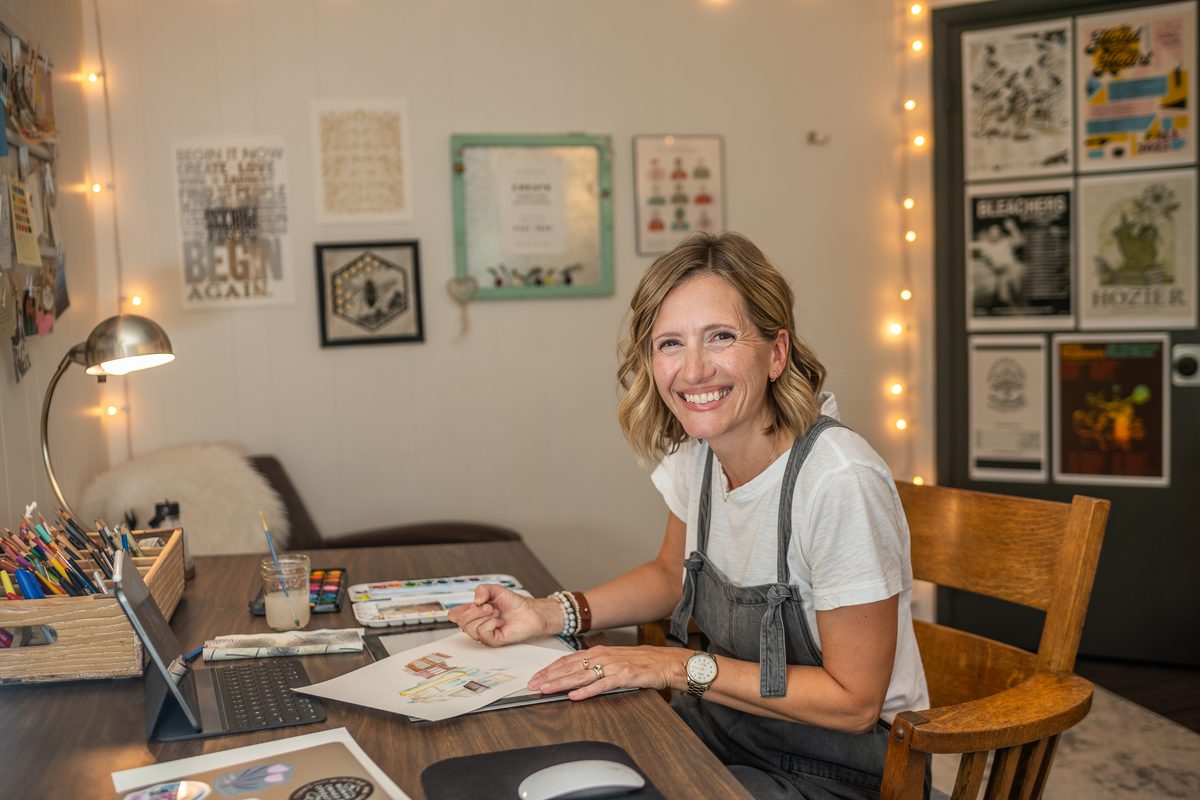A smiling artist at her desk painting a watercolor, with brushes, paints and framed prints around her.