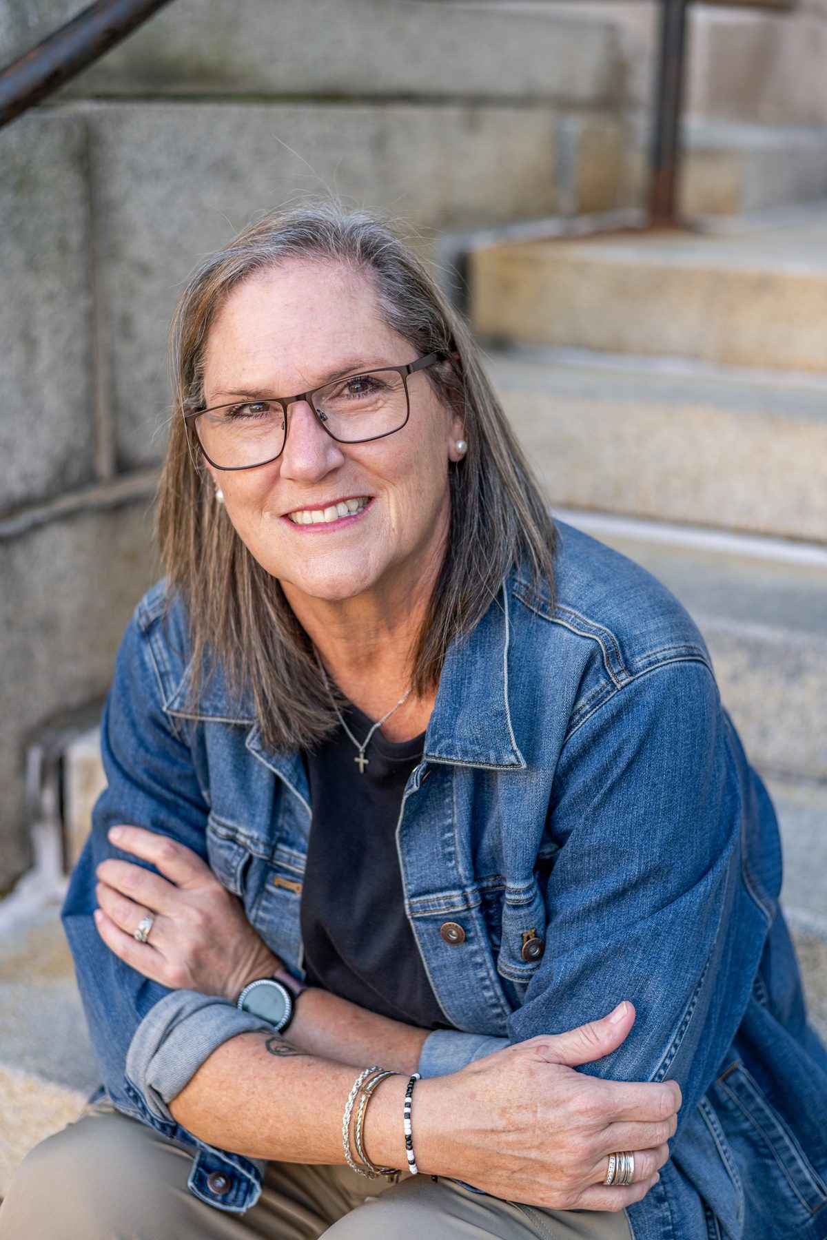 A smiling woman with glasses and shoulder-length hair, in a denim jacket, seated on stone steps.