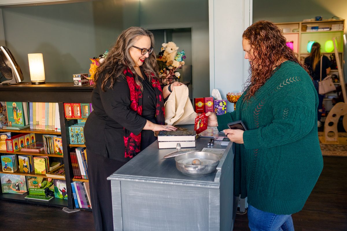 Two women at a shop counter as the owner, smiling in a red floral scarf, rings up a customer who is paying with her phone.