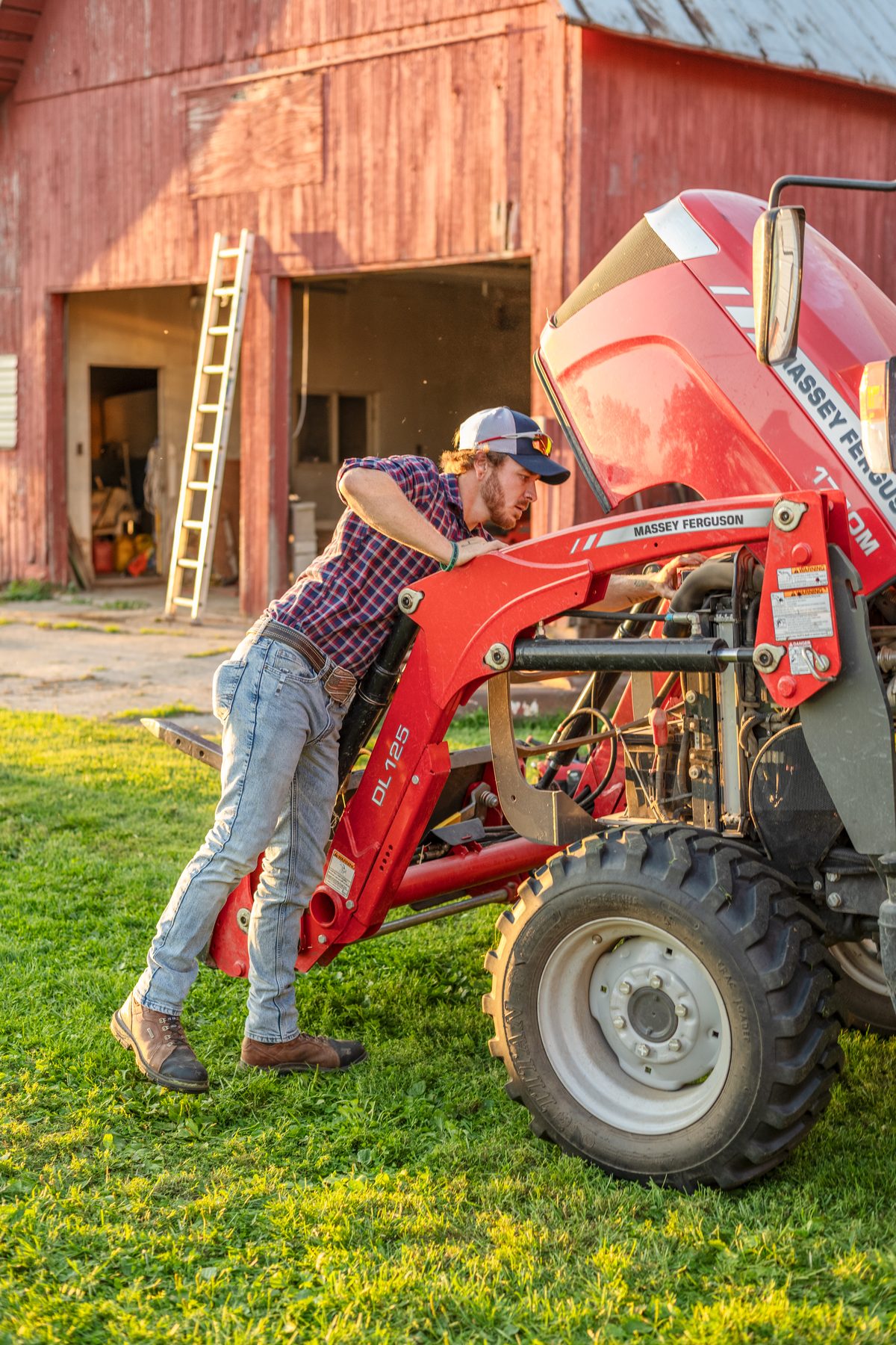 A man in a plaid shirt leans over the open hood of a red tractor on a sunlit lawn in front of a red barn with a ladder.