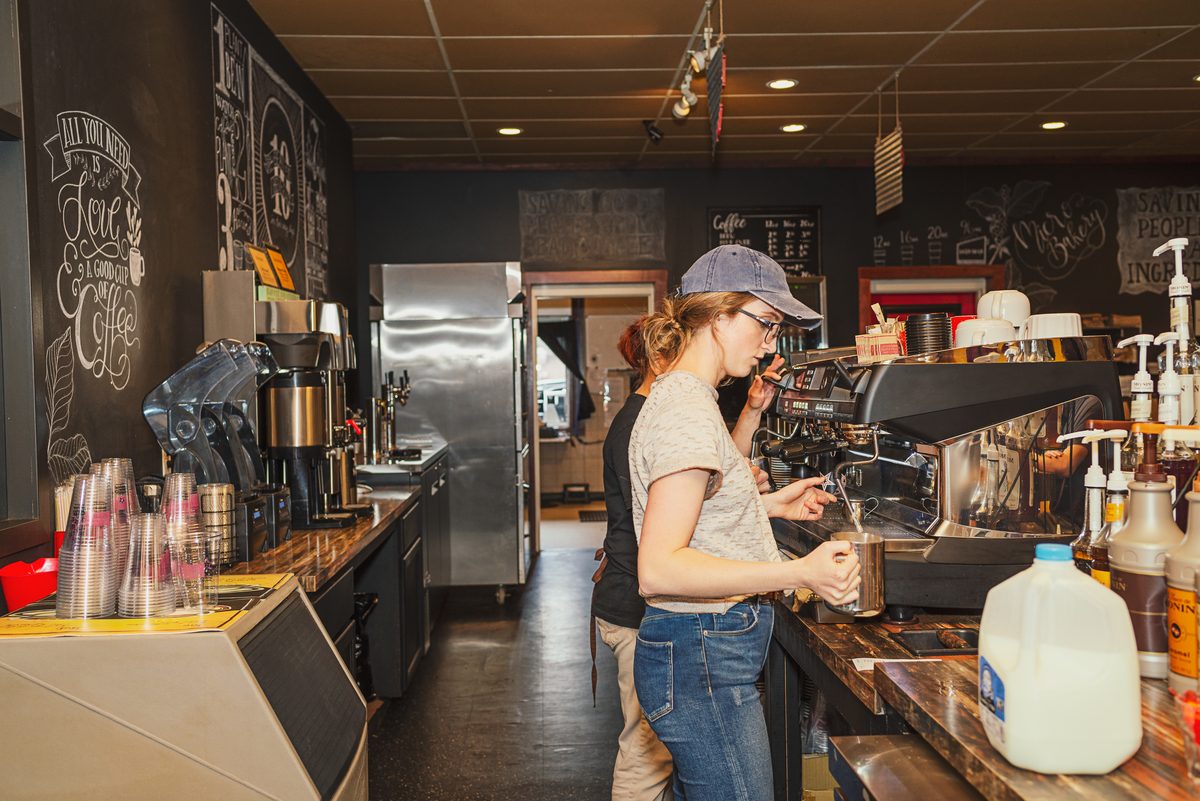 A barista in a ball cap works the espresso machine behind a coffee shop counter, with chalkboard walls, cups and a jug of milk around her.