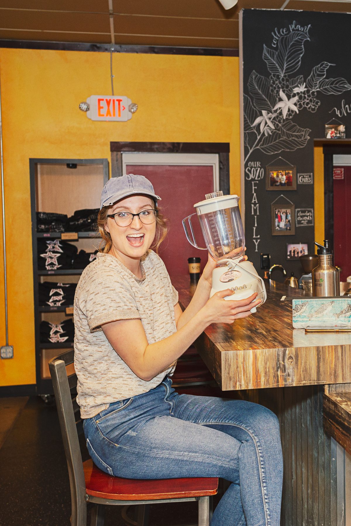 A smiling woman in a ball cap and glasses sits at a wooden counter holding a blender, in a coffee shop with a yellow wall and a chalkboard behind her.