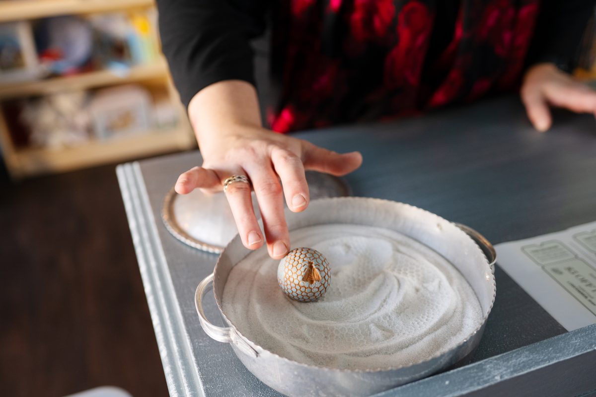 A hand gently rolls a patterned egg through a tray of white sand on a gray-blue table.