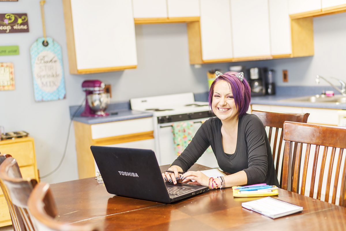 A smiling woman with purple hair works on a laptop at a wooden dining table in a bright kitchen, with a notebook and highlighters beside her.